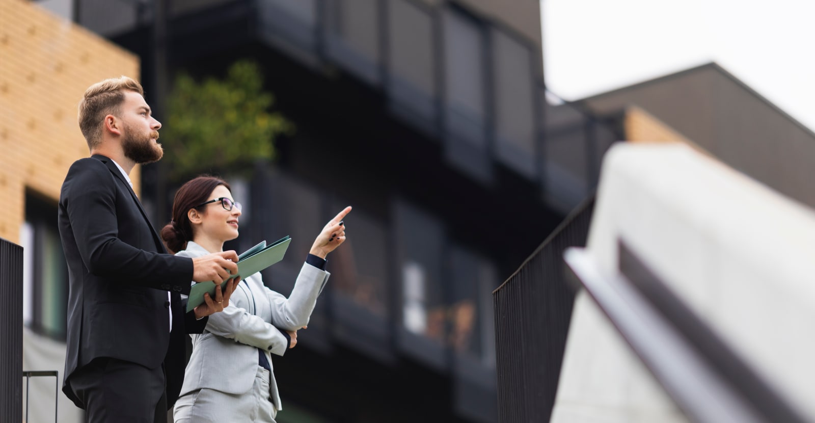 A man and a woman in business outfits are standing outside a building. The woman is pointing to something.