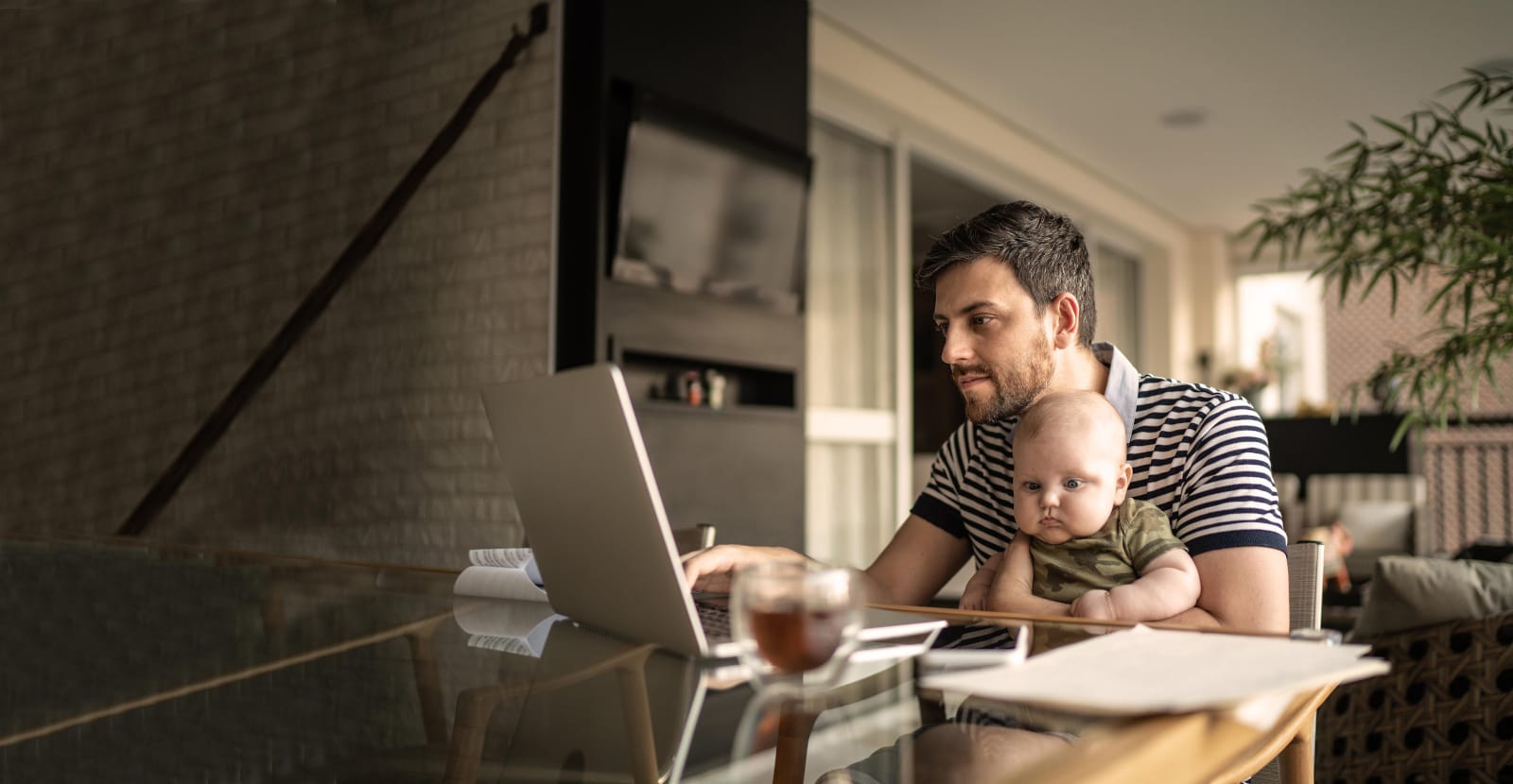 A father is sitting at his laptop, holding a child in his arms.