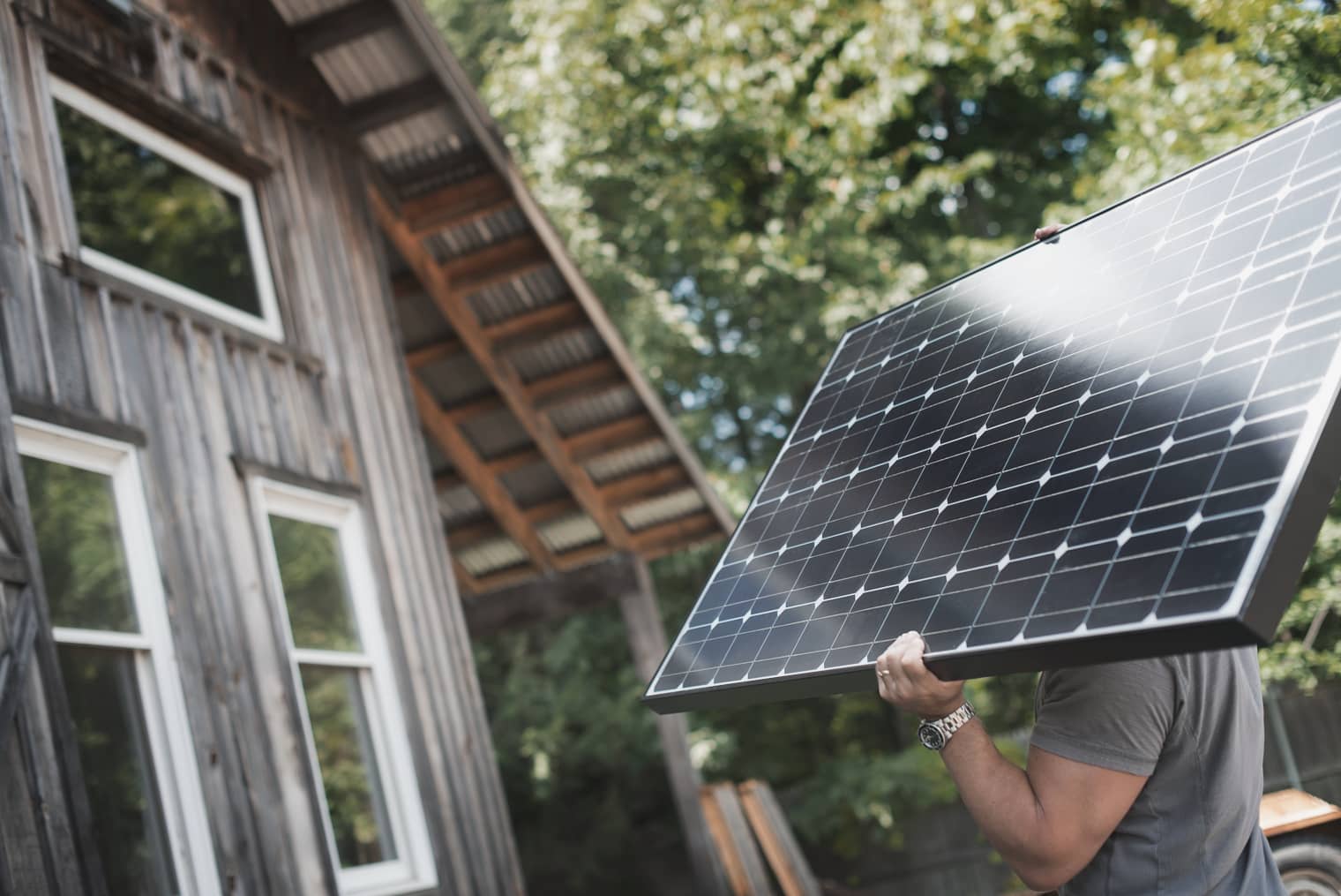 Ein Mann trägt ein Solarmodul zu einem Haus; im Hintergrund sind Bäume zu sehen.