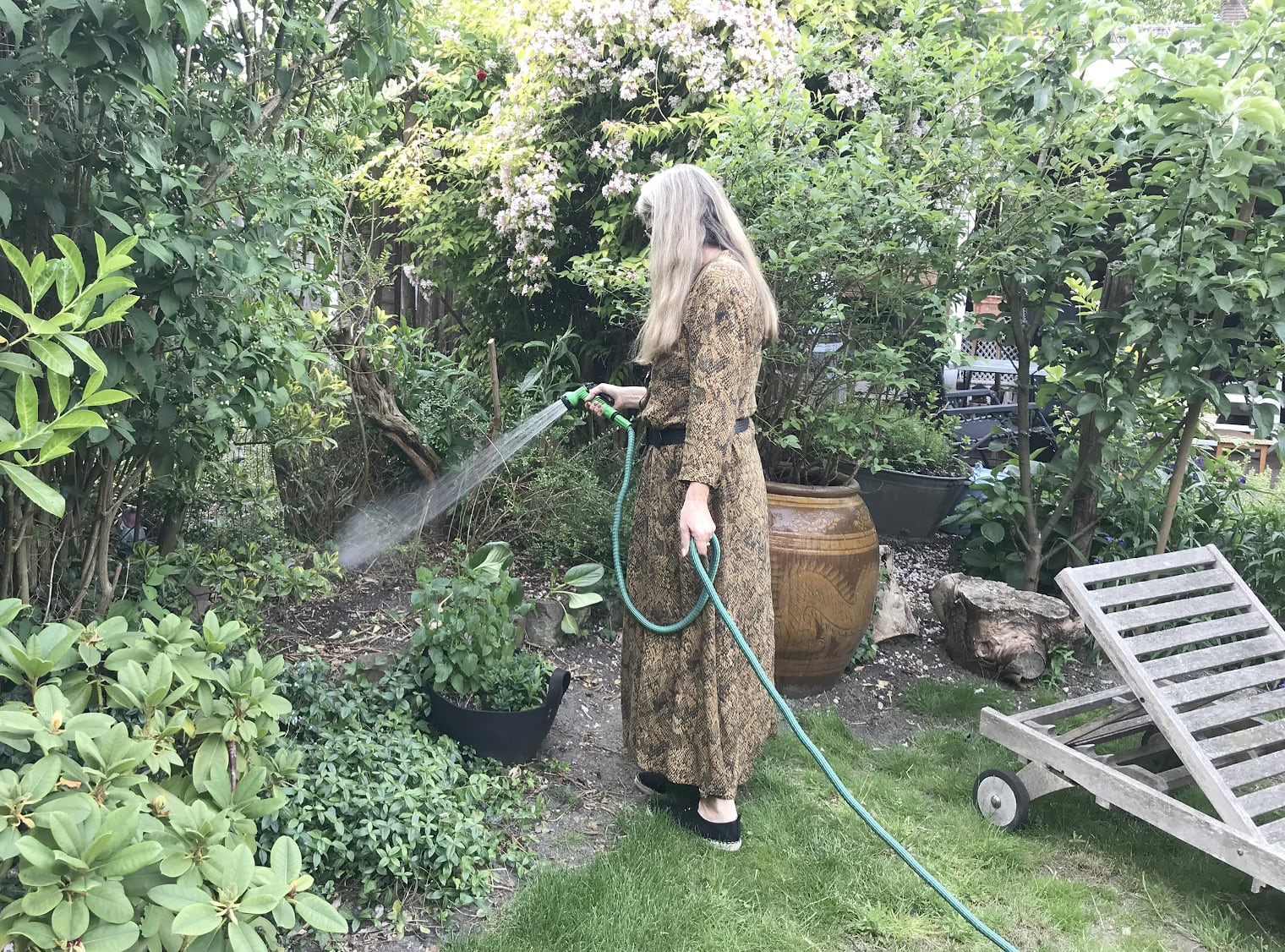 Eine ältere Frau mit grauem Haar bewässert den Garten ihres Einfamilienhauses.