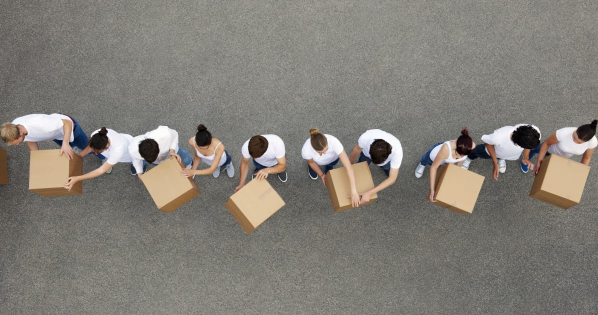 Ten people in white T-shirts can be seen from above, passing moving boxes to each other.